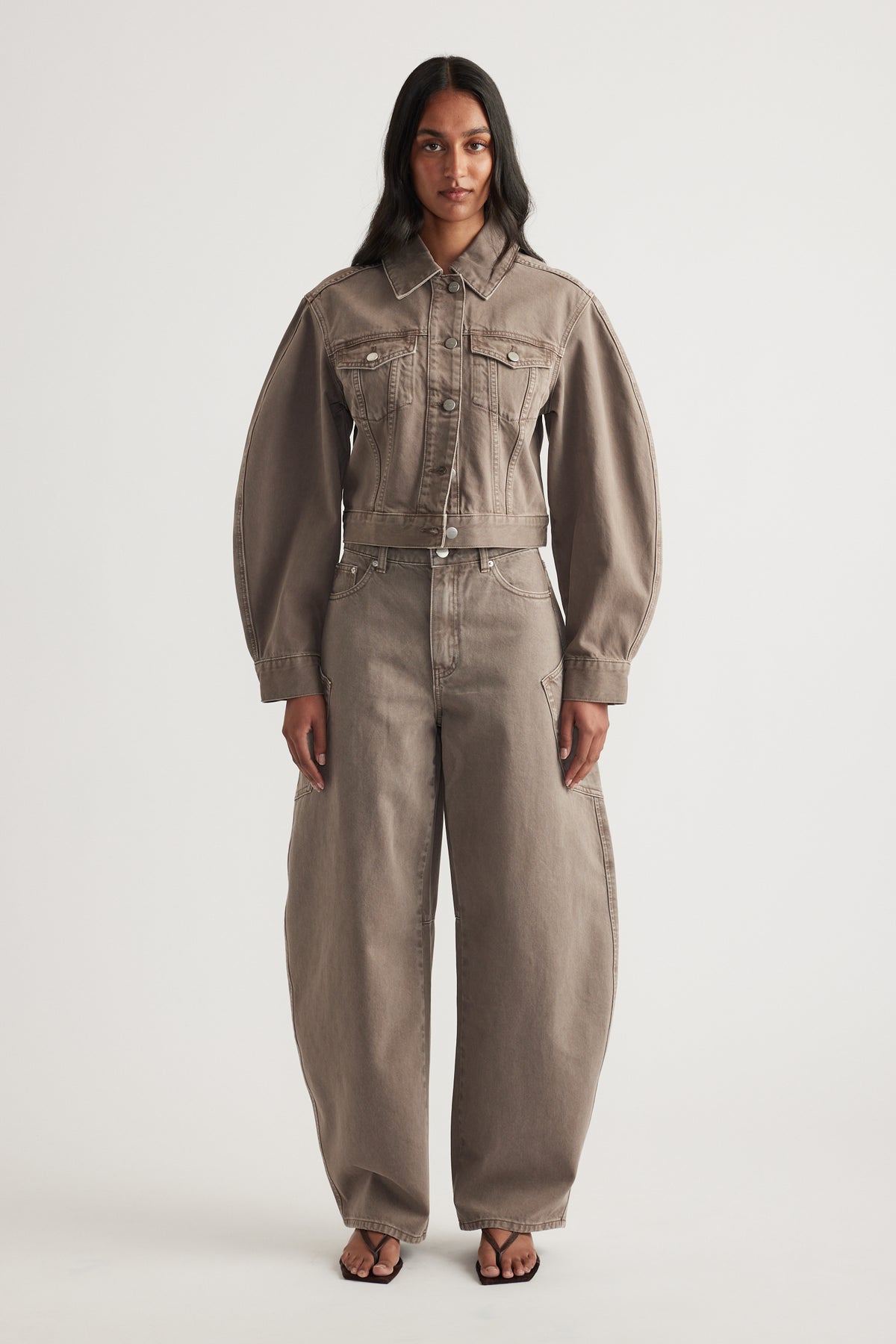 Model in a cropped taupe denim jacket with chest pockets, matching barrel leg jeans and black sandals