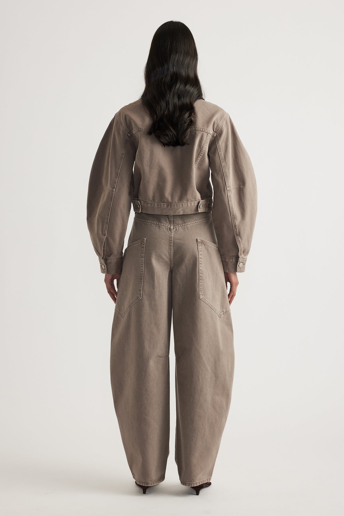 Woman seen from behind wearing a taupe cropped denim jacket with long sleeves and a cinched waist