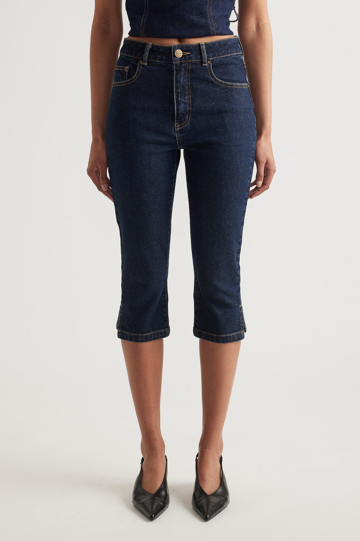 Woman wearing high rise dark wash denim capri pants with a slim fit and black pointed heels