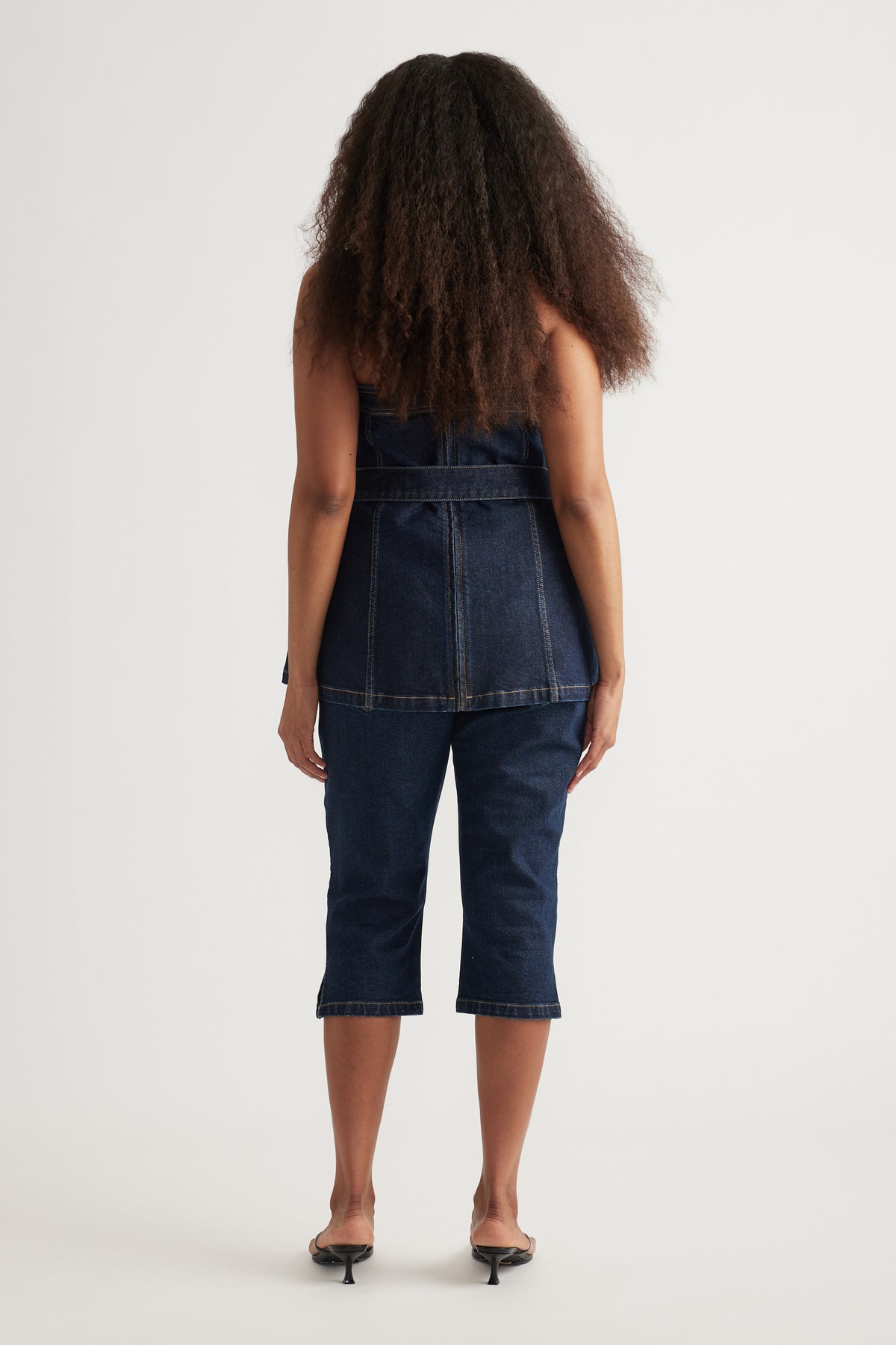 Back view of a model in a dark denim strapless longline top with matching capri jeans and heels