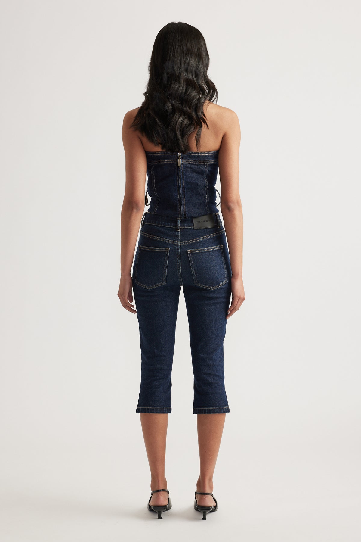 Back view of a woman wearing a strapless denim top with matching dark denim capri jeans and heels