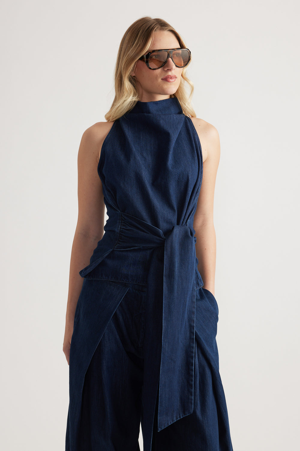 Woman wearing a navy sleeveless funnel neck top with a waist tie and oversized sunglasses