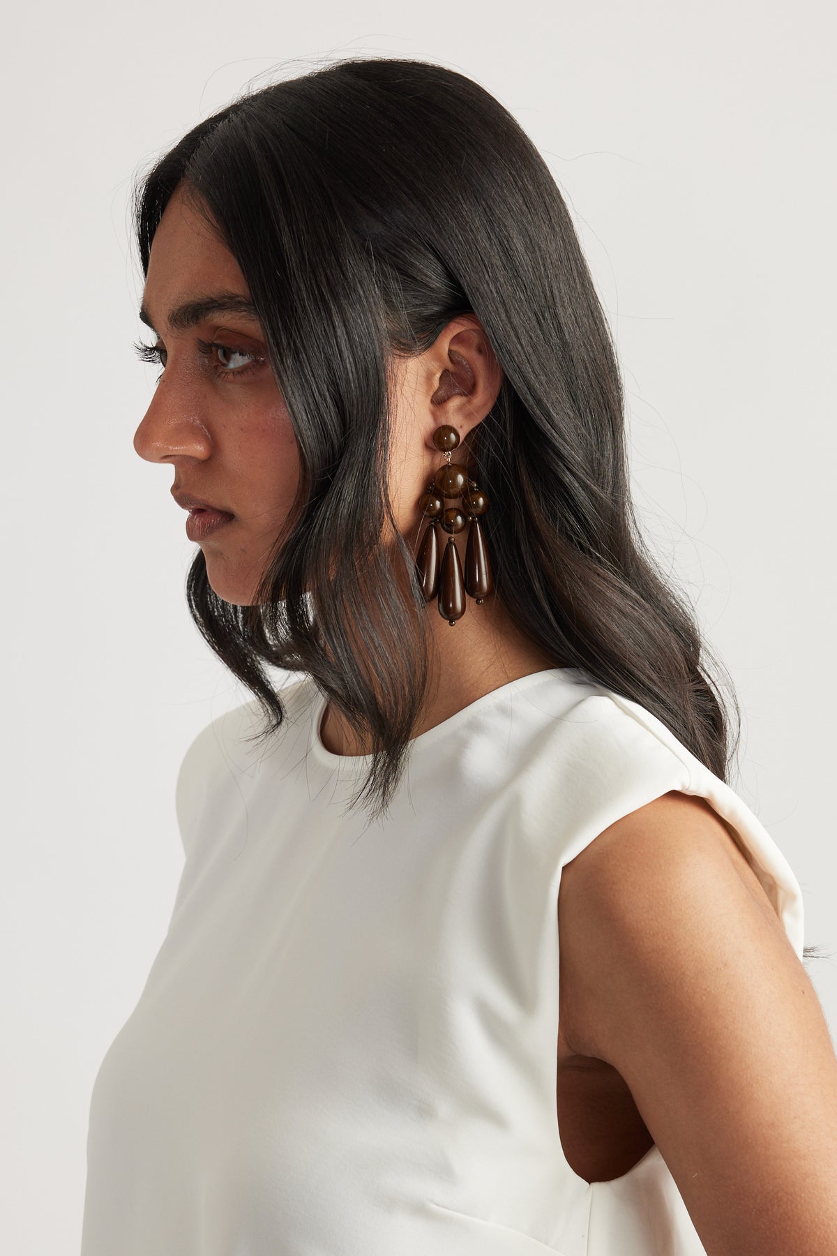 Profile of woman with dark hair wearing statement brown beaded earrings and a sleeveless white top against a plain background