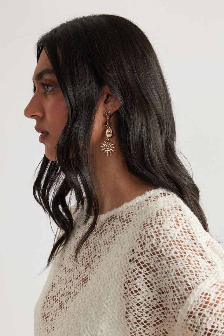 Profile of woman with dark hair wearing textured gold drop earrings featuring sun and pendant motifs and a cream knit top