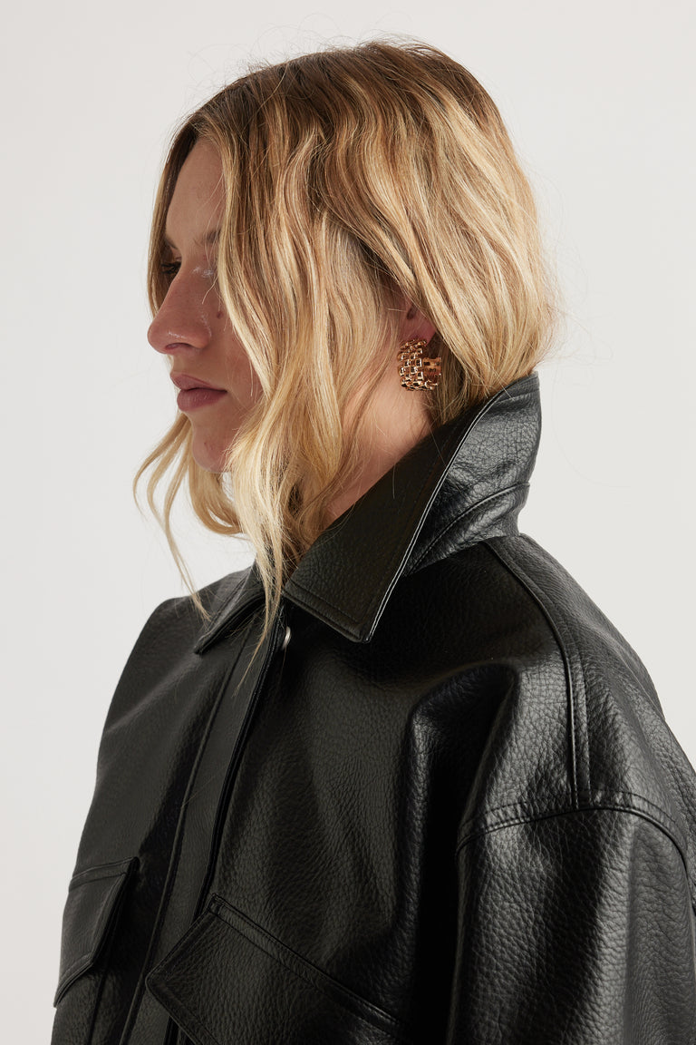 Woman with wavy blonde hair wearing textured black leather jacket and chunky woven gold hoop earrings seen in profile