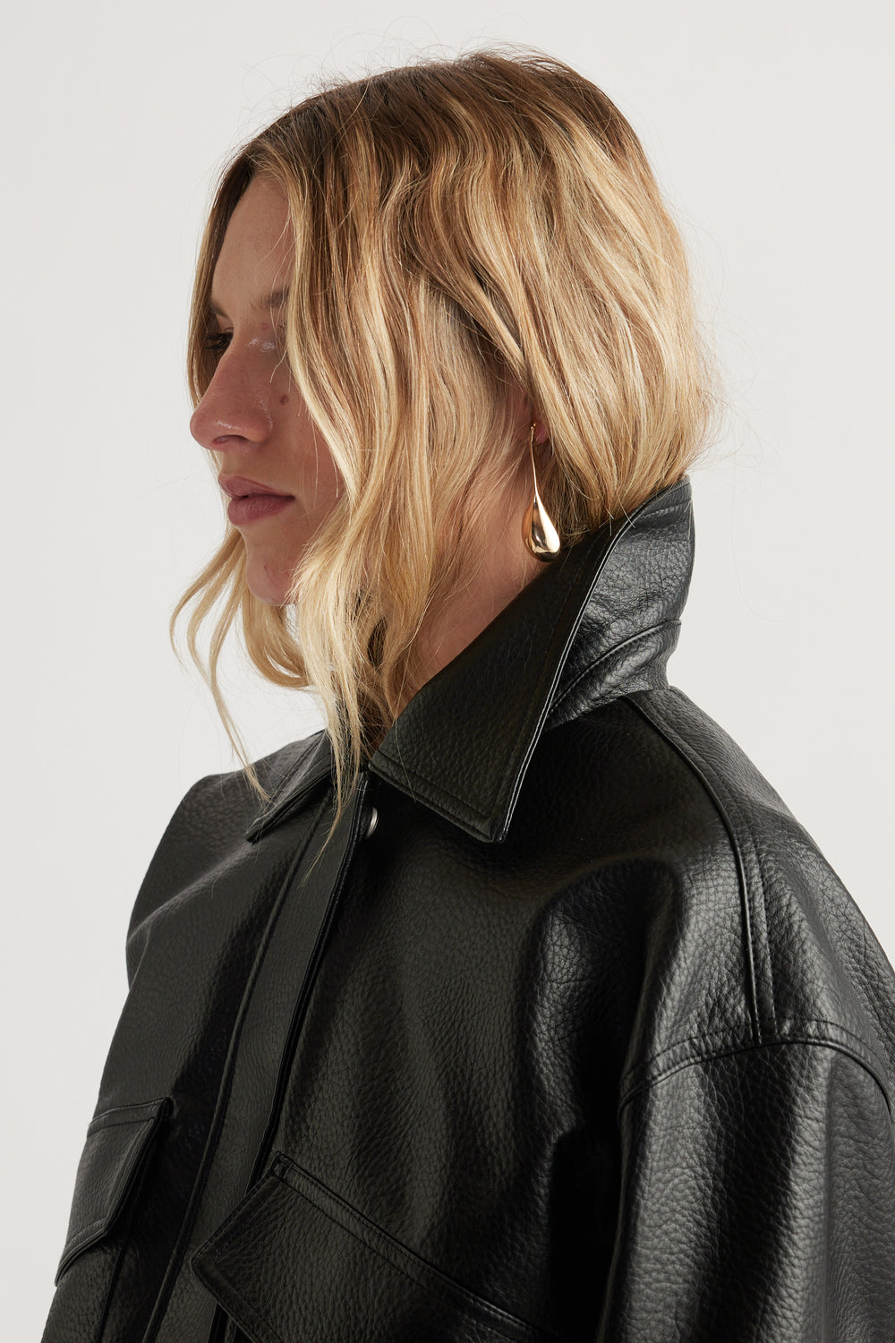 Profile of woman with wavy blonde hair wearing shiny gold teardrop earrings and a black textured jacket with wide collar