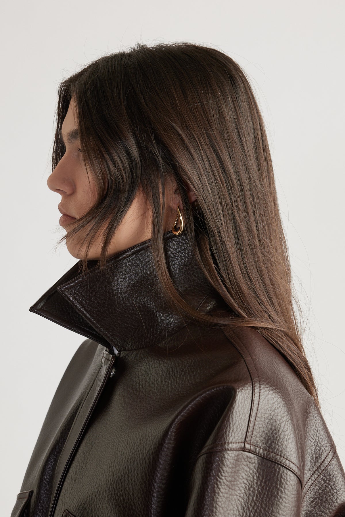 Side profile of a woman wearing small gold curved hoop earrings paired with a dark brown textured leather jacket.