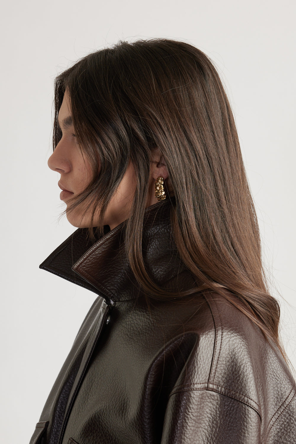 Profile of woman with long brown hair wearing textured dark brown leather jacket and gold crinkle hoop earrings