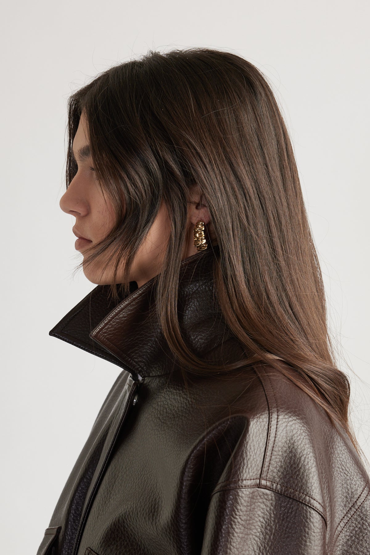 Profile of woman with long brown hair wearing textured dark brown leather jacket and gold crinkle hoop earrings