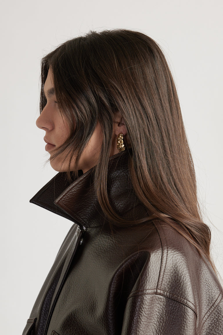 Profile of woman with long brown hair wearing textured dark brown leather jacket and gold crinkle hoop earrings