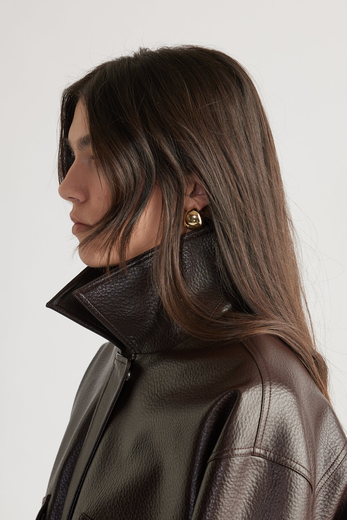 Side profile of a woman with long dark hair wearing bold gold hoop earrings and a dark textured leather jacket with a raised collar
