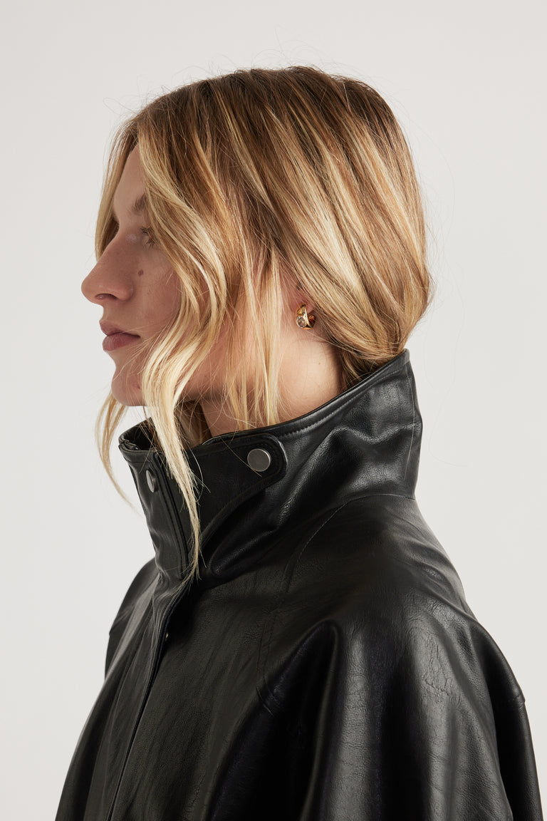Woman with loose blonde hair wearing a black leather jacket and small gold chunky hoop earrings in profile view