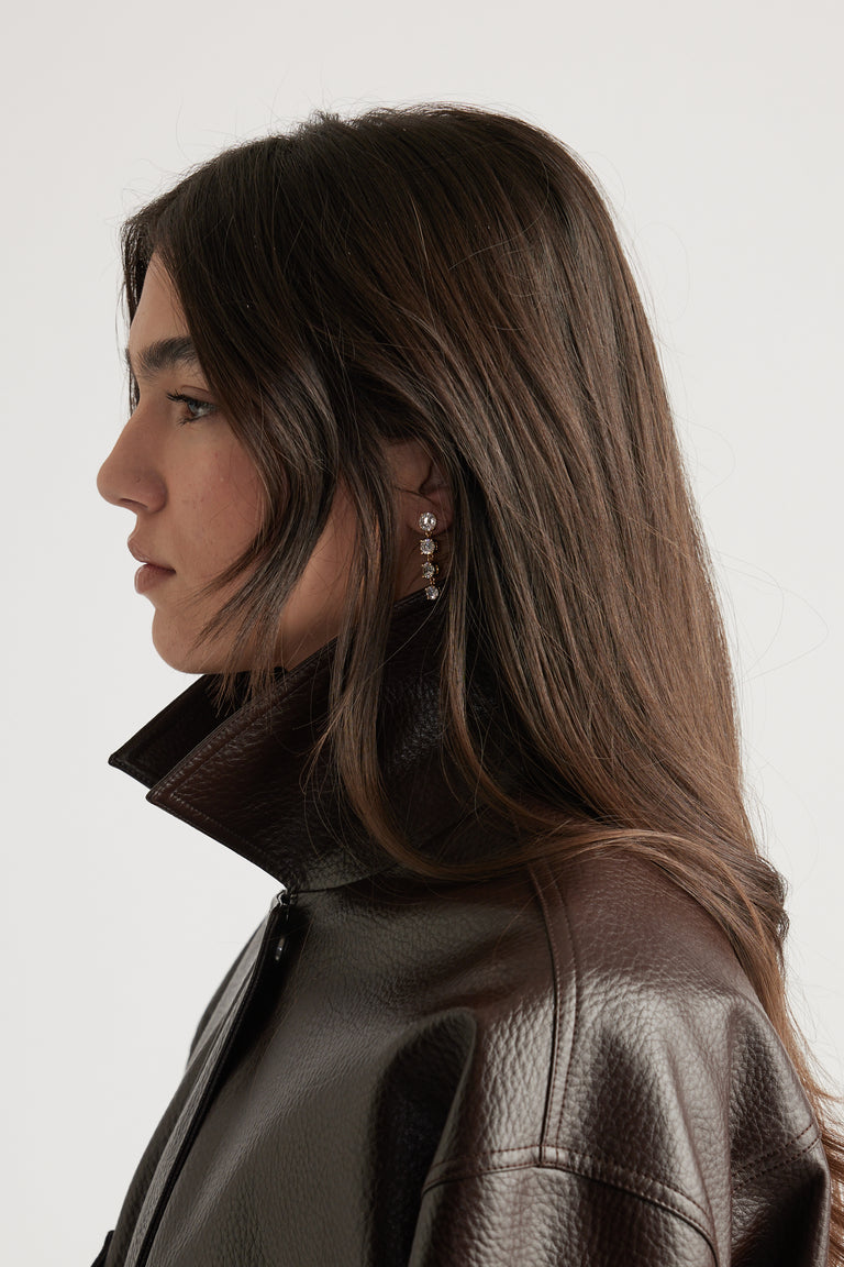 Profile of woman wearing glossy dark brown leather jacket and delicate gold-tone drop earrings with sparkling stones