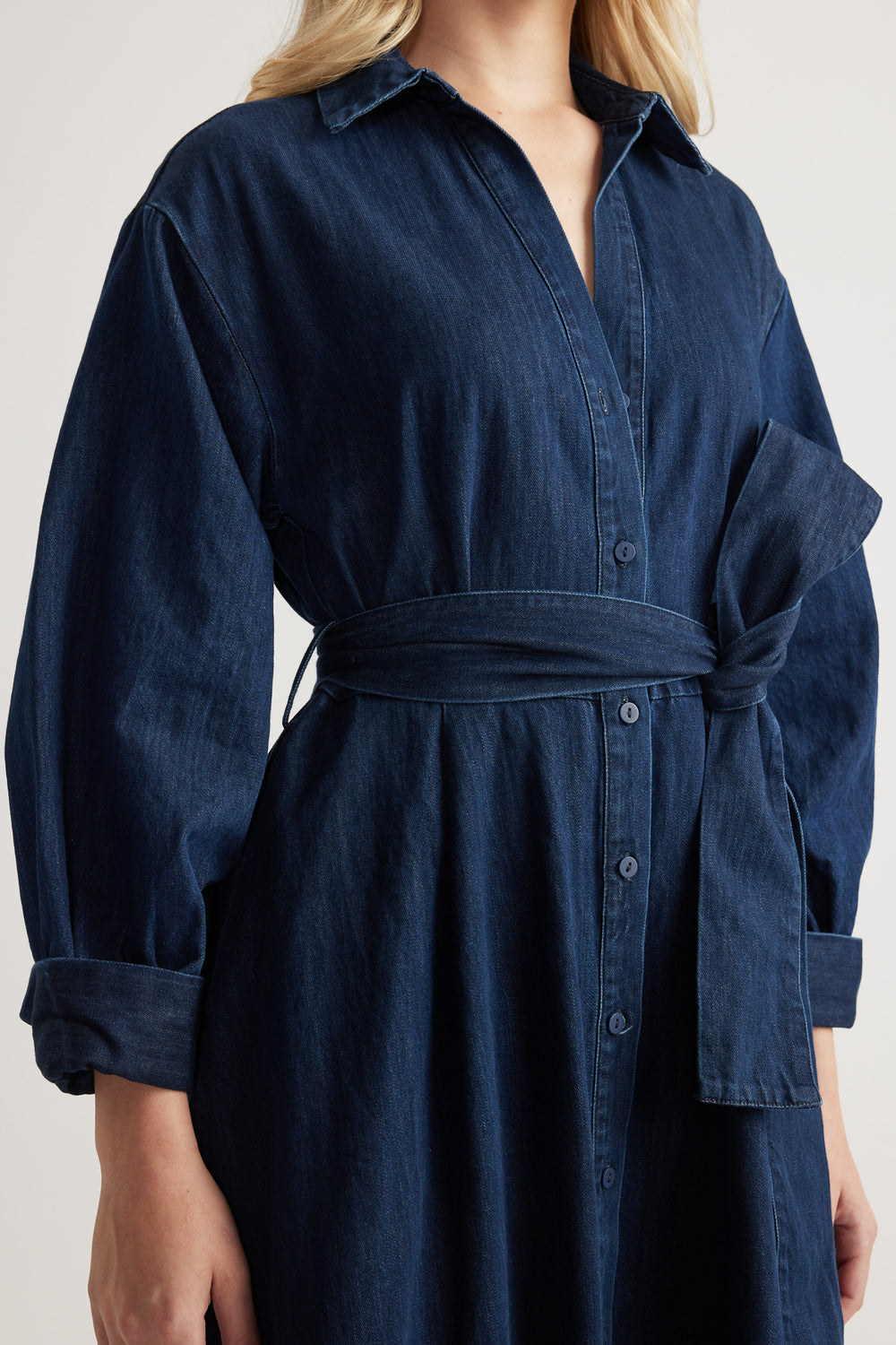 Woman wearing a dark denim midi shirt dress with long sleeves, collar and adjustable waist tie