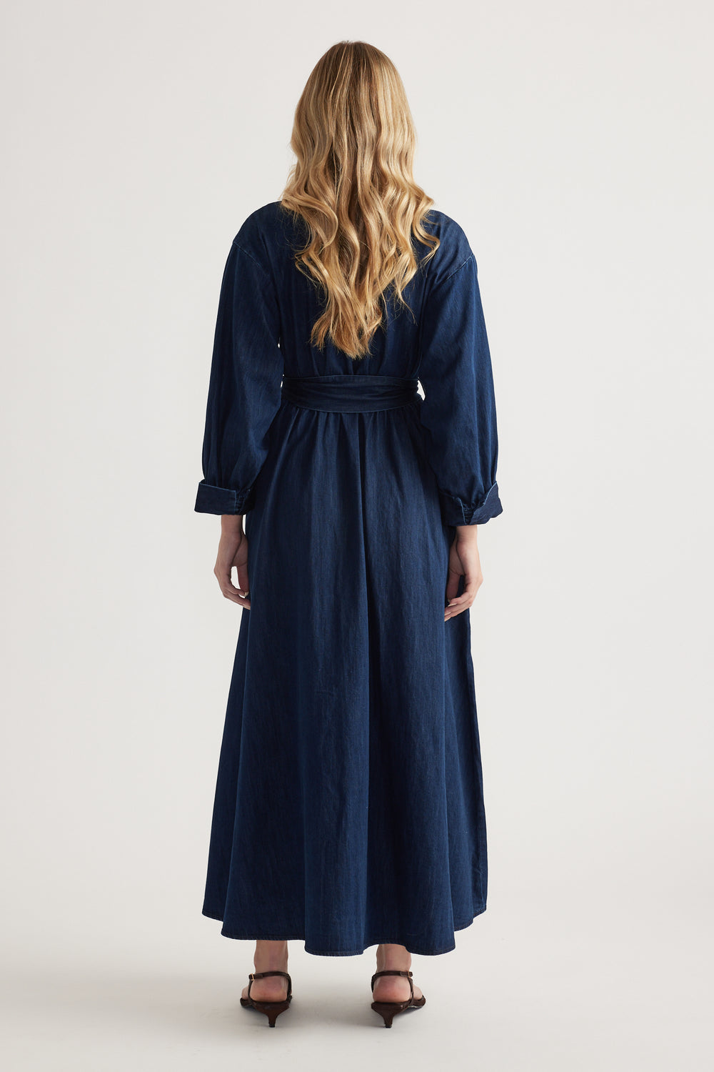 Back view of a woman wearing a dark denim midi shirt dress with long sleeves and a tied waist