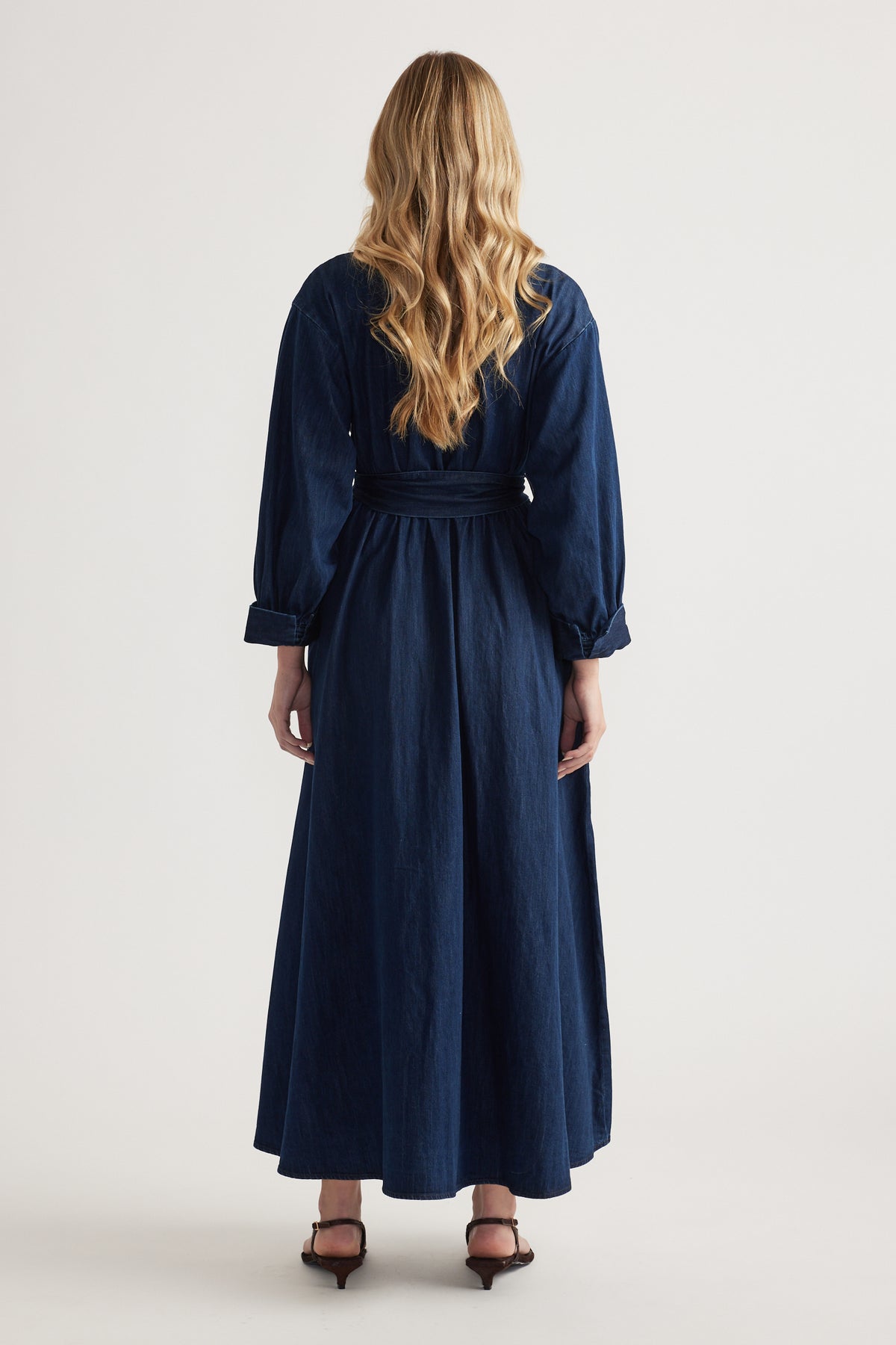 Back view of a woman wearing a dark denim midi shirt dress with long sleeves and a tied waist