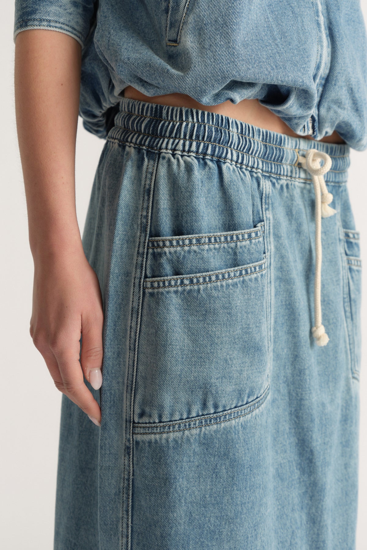 Close-up of a model wearing a light wash denim midi skirt with an elastic waist, drawstring and patch pockets
