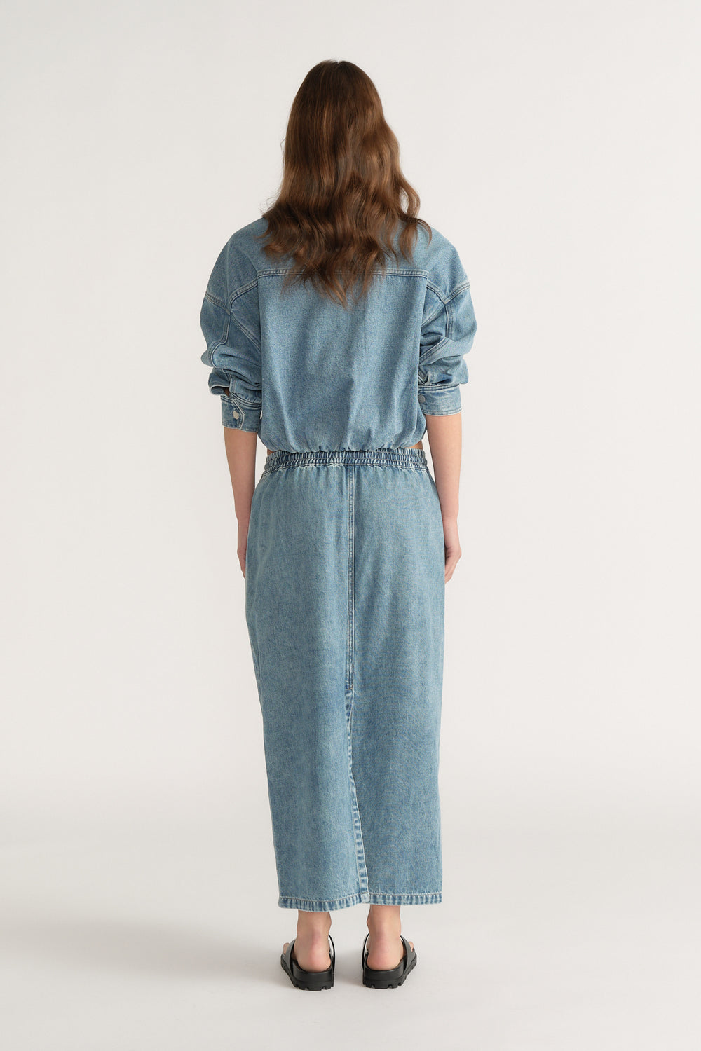 Back view of a woman wearing a denim midi skirt with an elastic waist and matching denim jacket