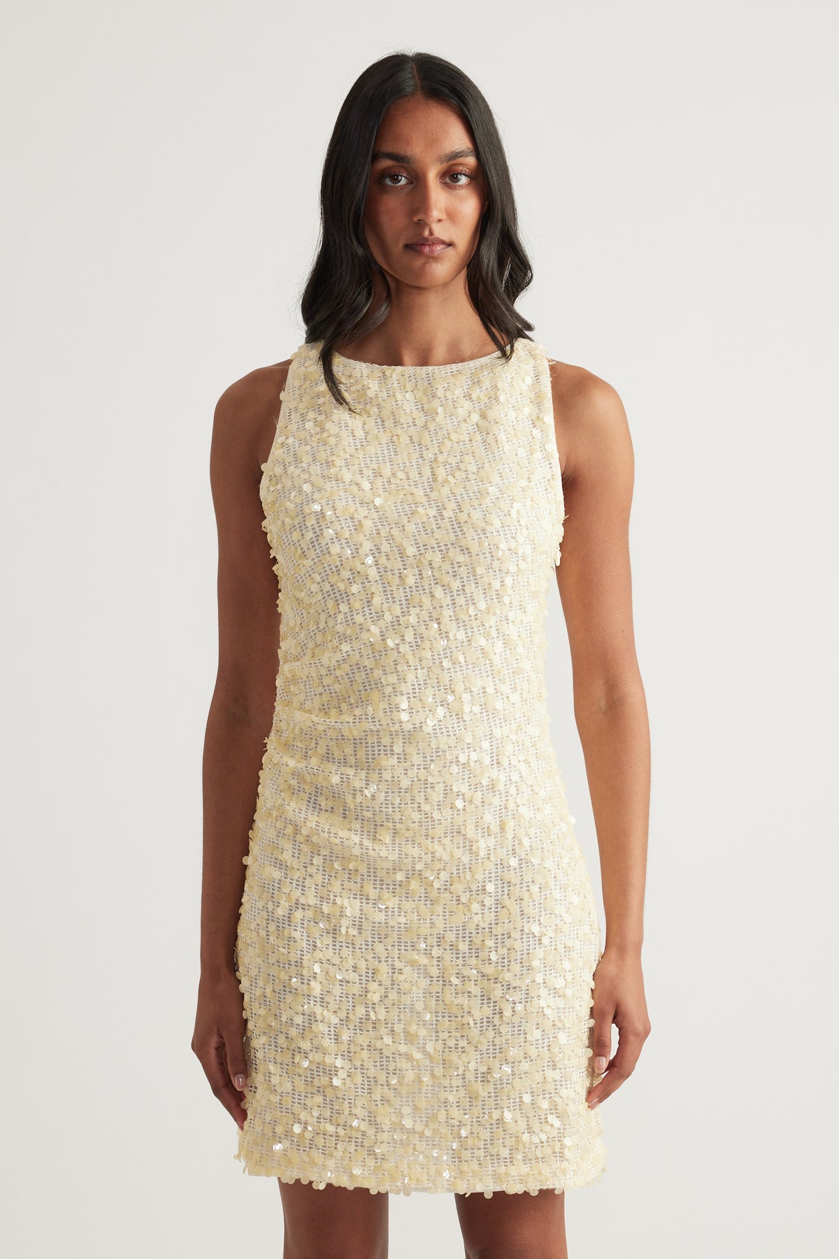 Model wearing a cream sequin mini dress with a sleeveless boat neckline, scoop back and fitted silhouette