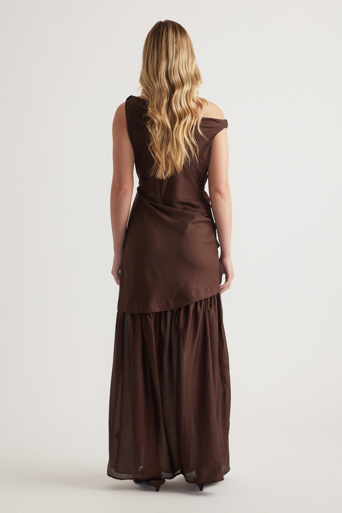 Woman wearing a dark brown draped maxi dress with an off shoulder neckline, viewed from behind