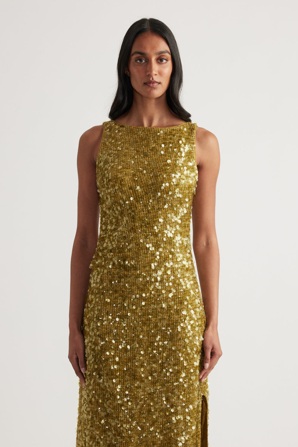 Woman wearing a fitted gold sequin maxi dress with a sleeveless boat neckline and side slit