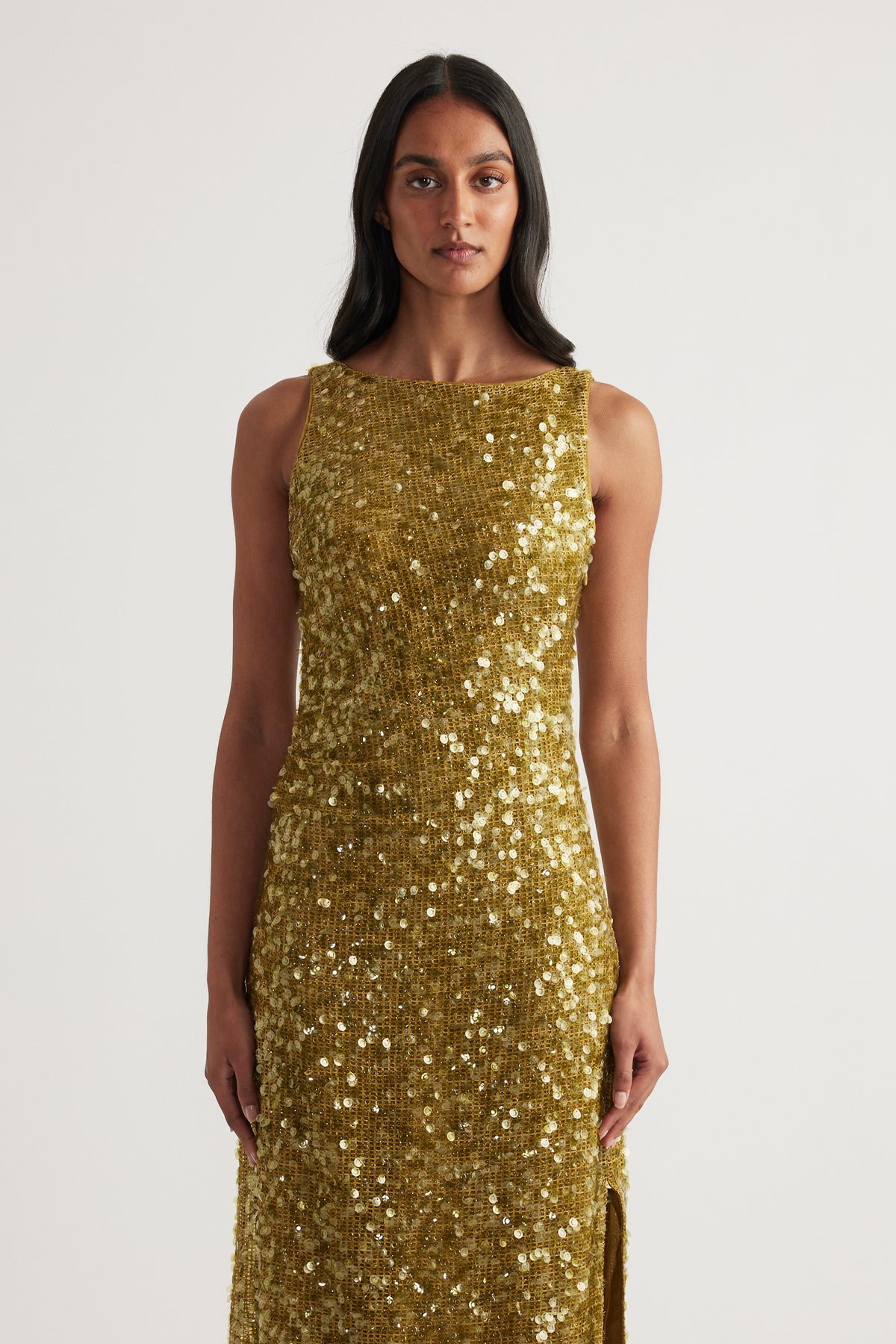 Woman wearing a fitted gold sequin maxi dress with a sleeveless boat neckline and side slit