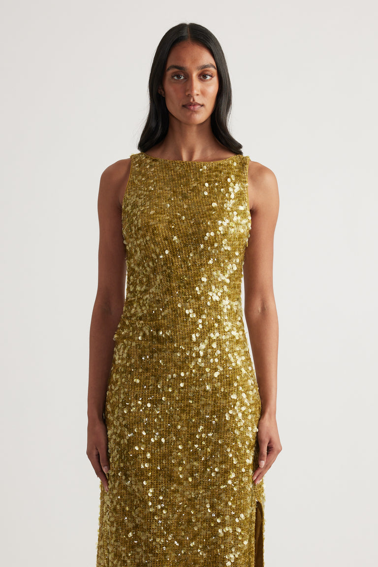 Woman wearing a fitted gold sequin maxi dress with a sleeveless boat neckline and side slit