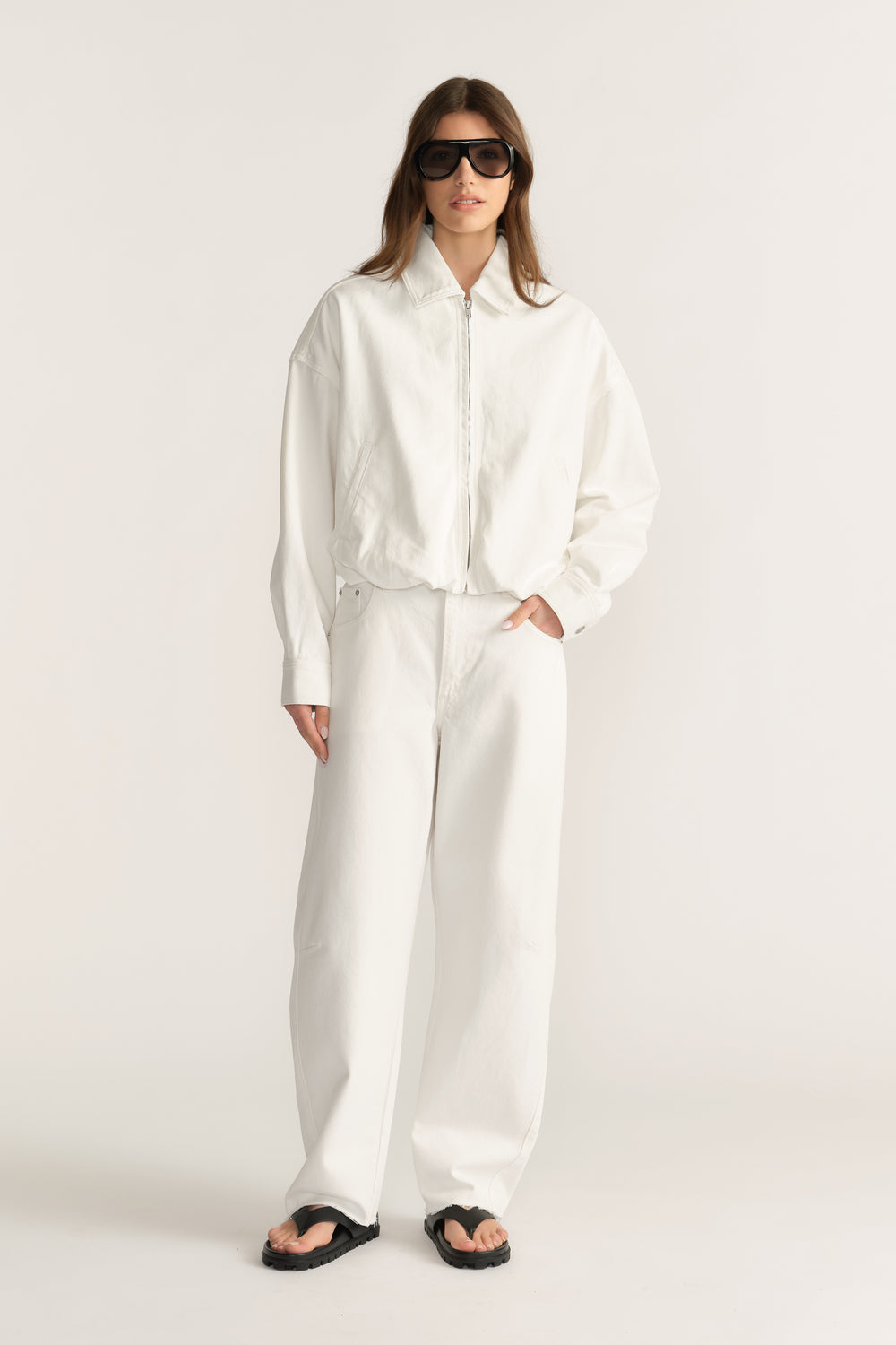 Woman wearing a white denim bomber jacket with wide leg pants, sunglasses and sandals