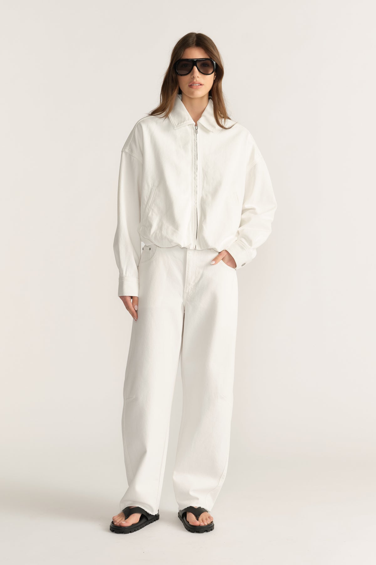 Woman wearing a white denim bomber jacket with wide leg pants, sunglasses and sandals