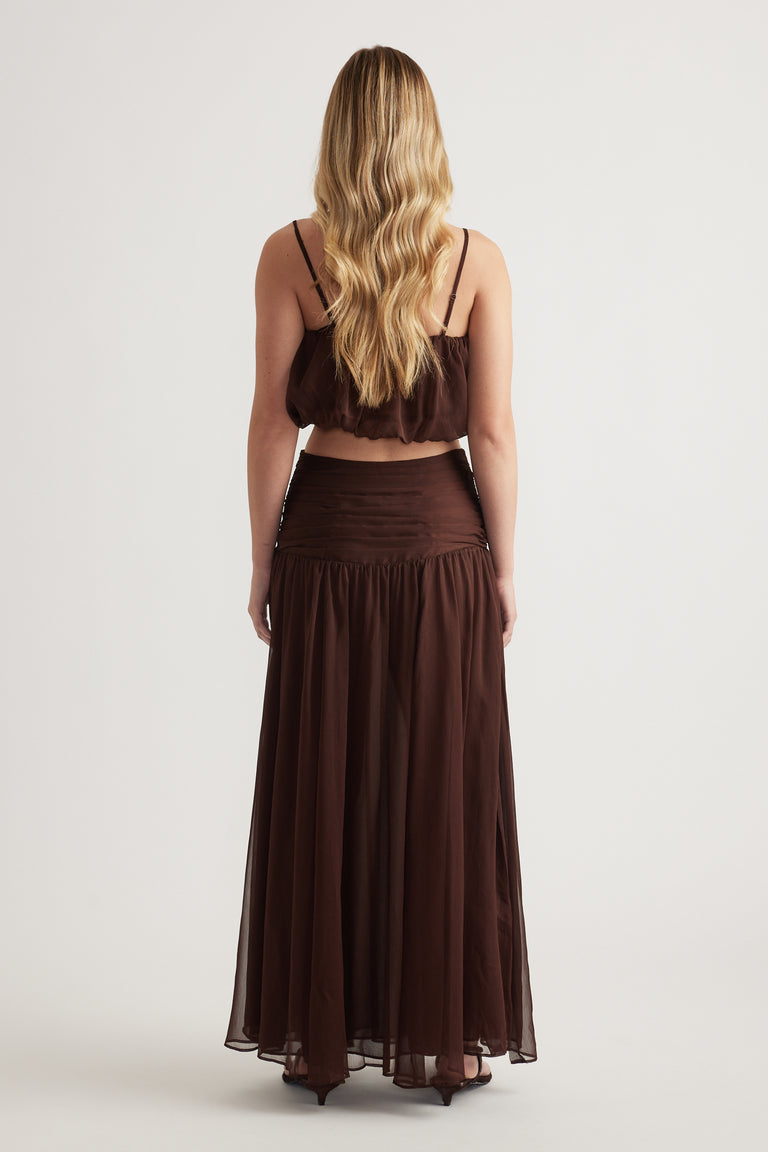 Back view of a model wearing a chocolate brown high waisted maxi skirt with gathered details and an ankle length hem