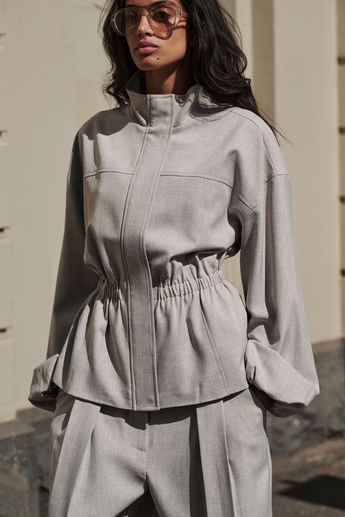 Woman wearing a grey cinched funnel neck jacket with matching wide leg pants and sunglasses