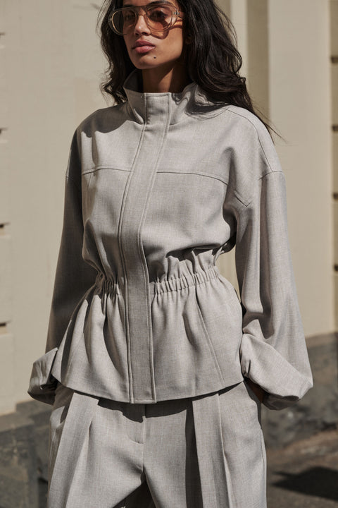 Woman wearing a grey cinched funnel neck jacket with matching wide leg pants and sunglasses