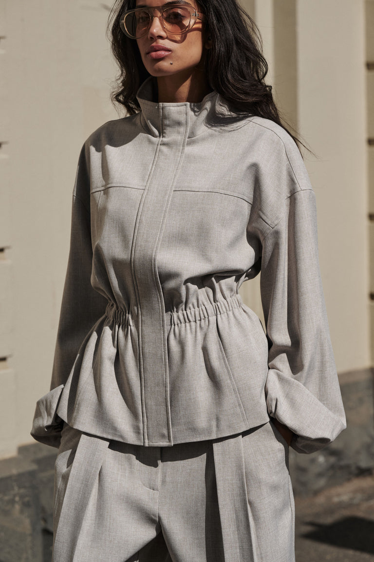 Woman wearing a grey cinched funnel neck jacket with matching wide leg pants and sunglasses