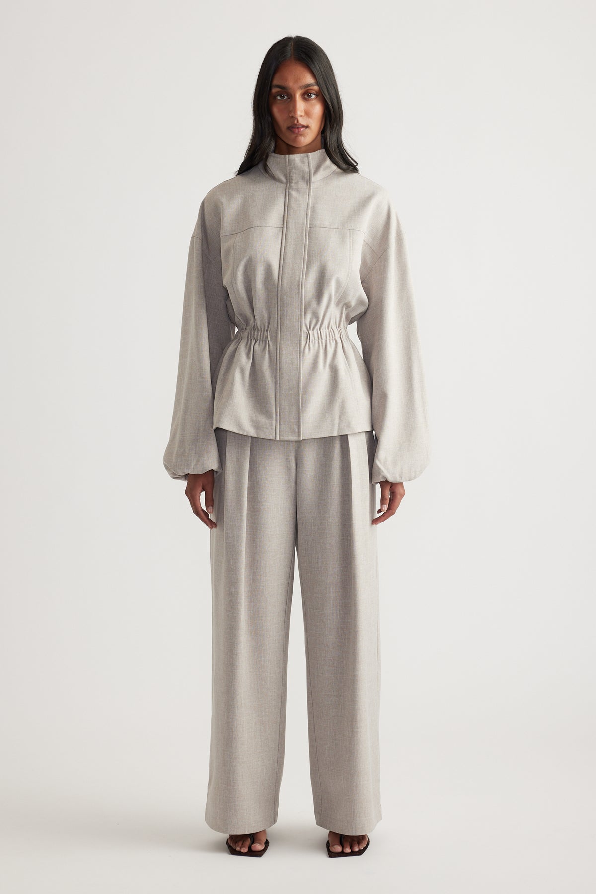 Woman wearing a taupe funnel neck jacket with a cinched waist, matching wide leg pants and black sandals