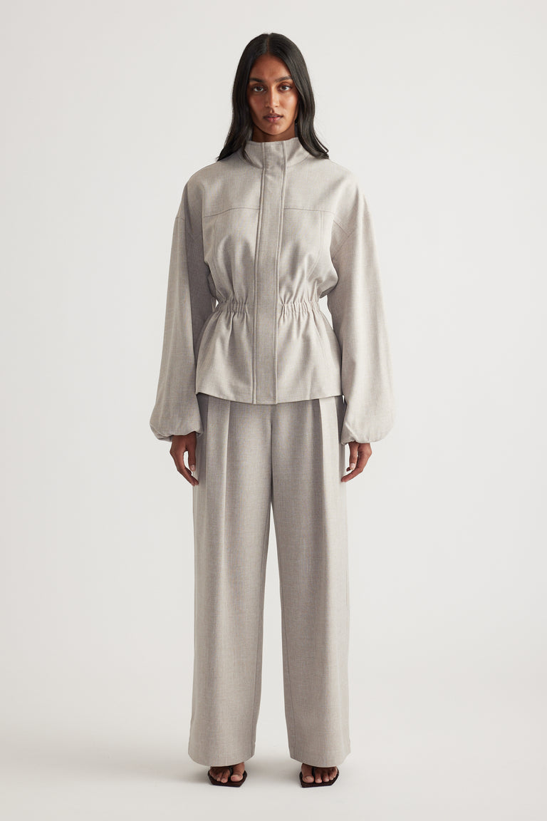 Woman wearing a taupe funnel neck jacket with a cinched waist, matching wide leg pants and black sandals