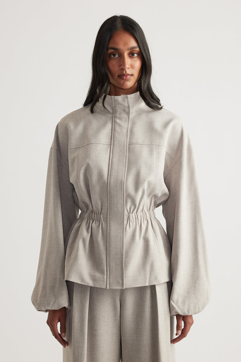 Woman wearing a taupe funnel neck cropped jacket with a cinched waist and matching wide leg pants