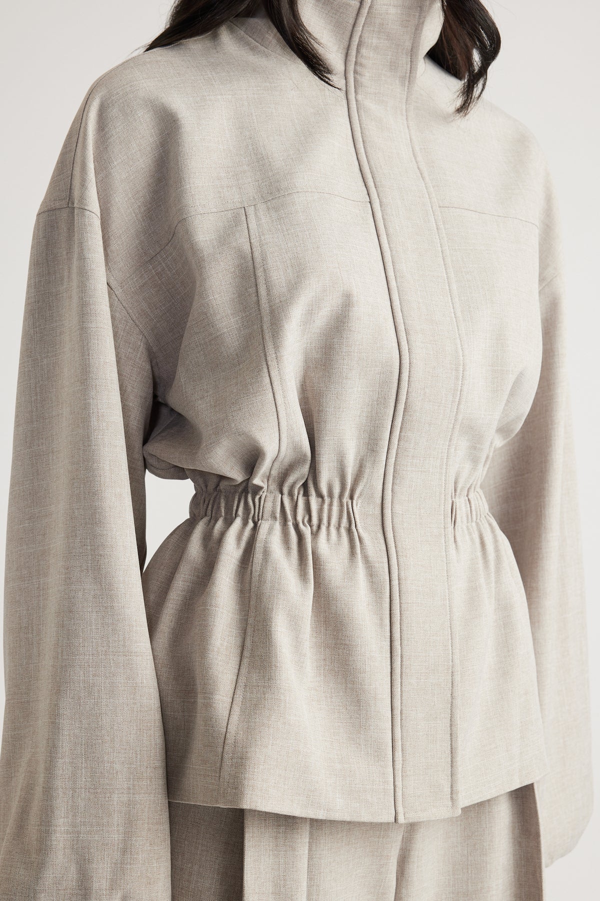Woman wearing a beige funnel neck jacket with a cinched waist and relaxed sleeves