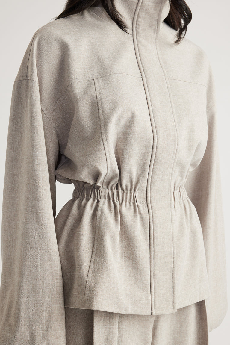 Woman wearing a beige funnel neck jacket with a cinched waist and relaxed sleeves