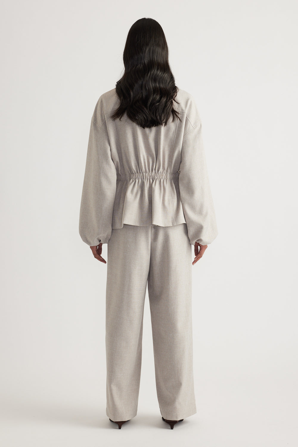 Back view of a woman wearing a beige cinched waist jacket with matching wide leg pants and black heels