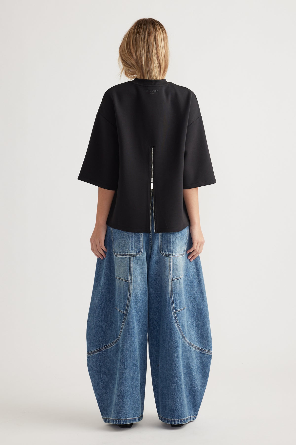 Woman wearing an oversized black top with back zipper detail paired with wide-leg patch pocket blue jeans viewed from behind
