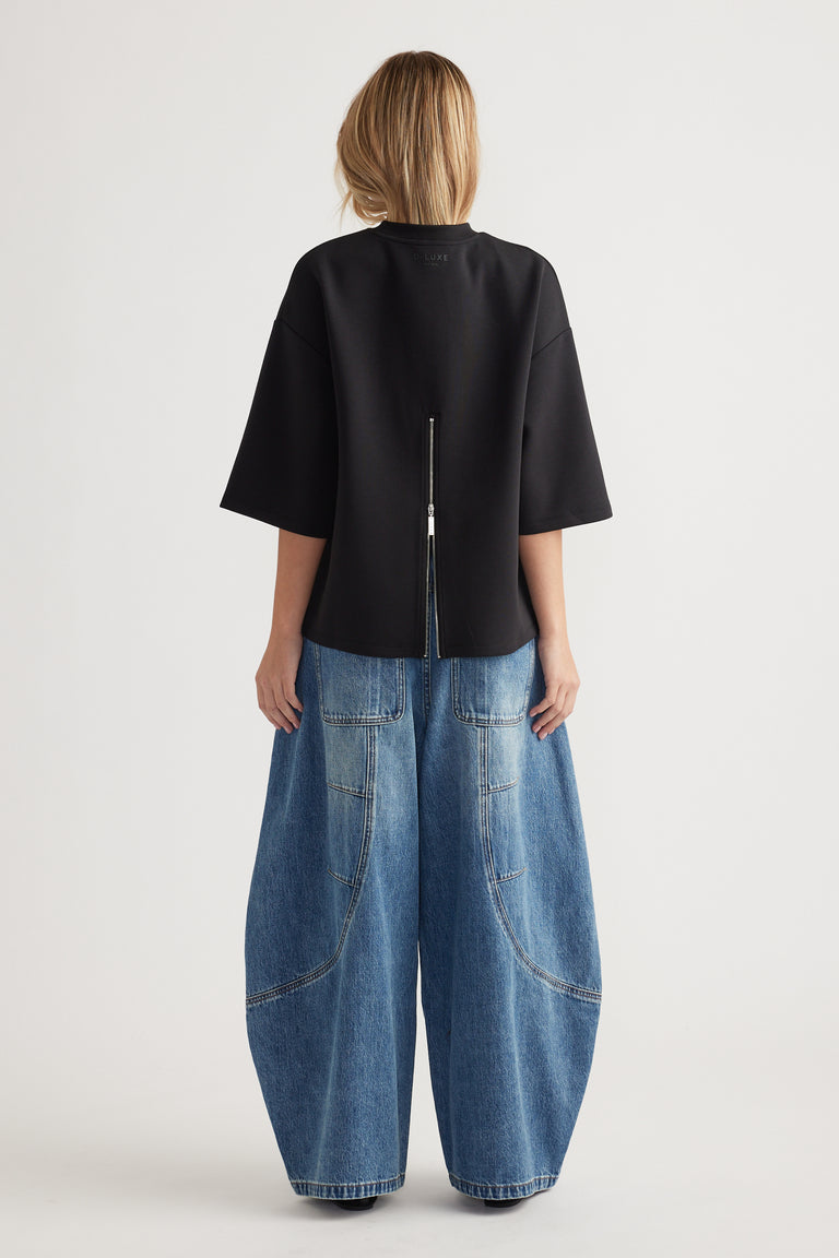 Woman wearing an oversized black top with back zipper detail paired with wide-leg patch pocket blue jeans viewed from behind