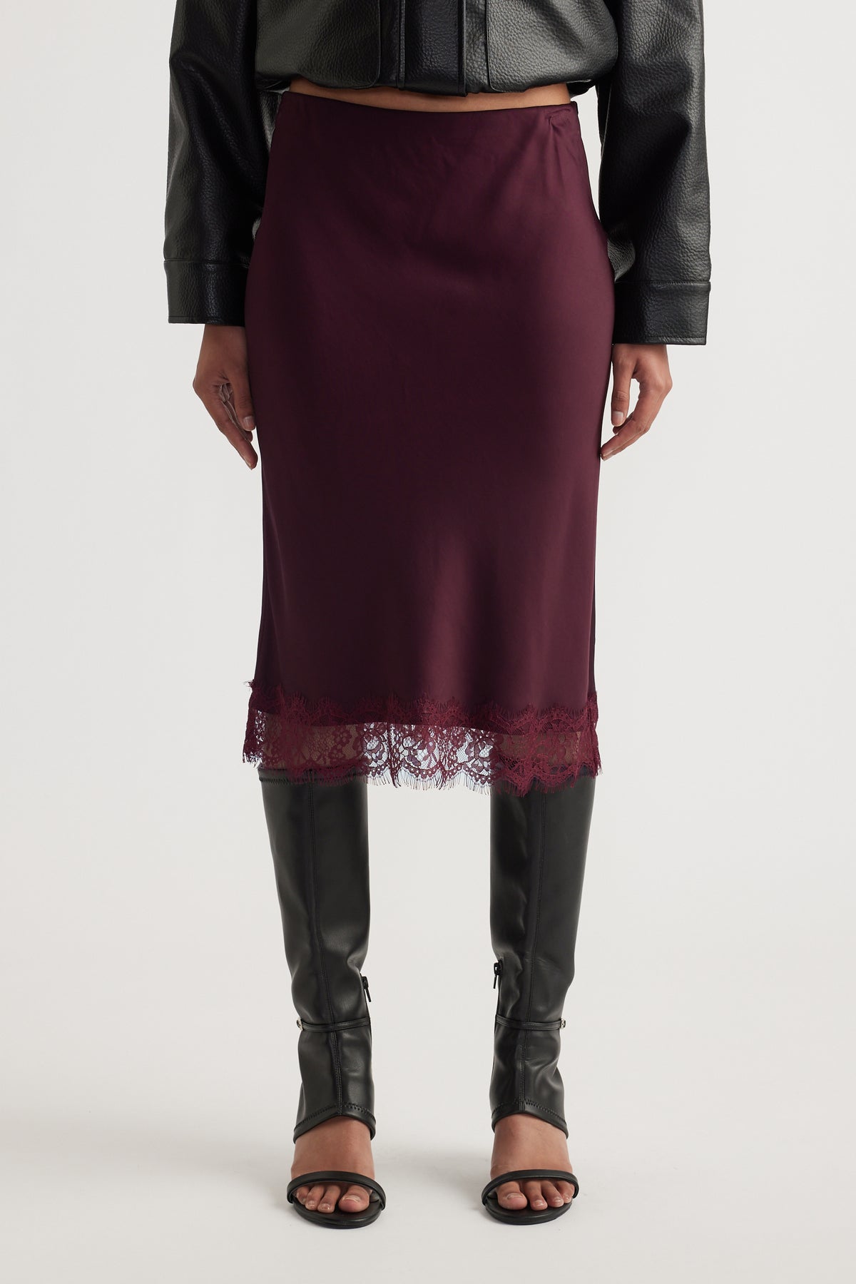 Woman wearing a burgundy satin midi skirt with lace trim, a black leather jacket and knee high boots