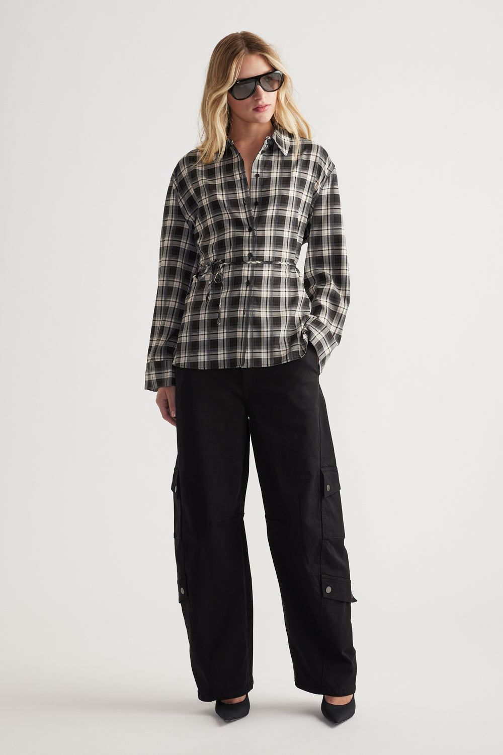 Woman wearing a relaxed fit black and white checked shirt with drawstring waist, paired with loose black cargo pants and sunglasses