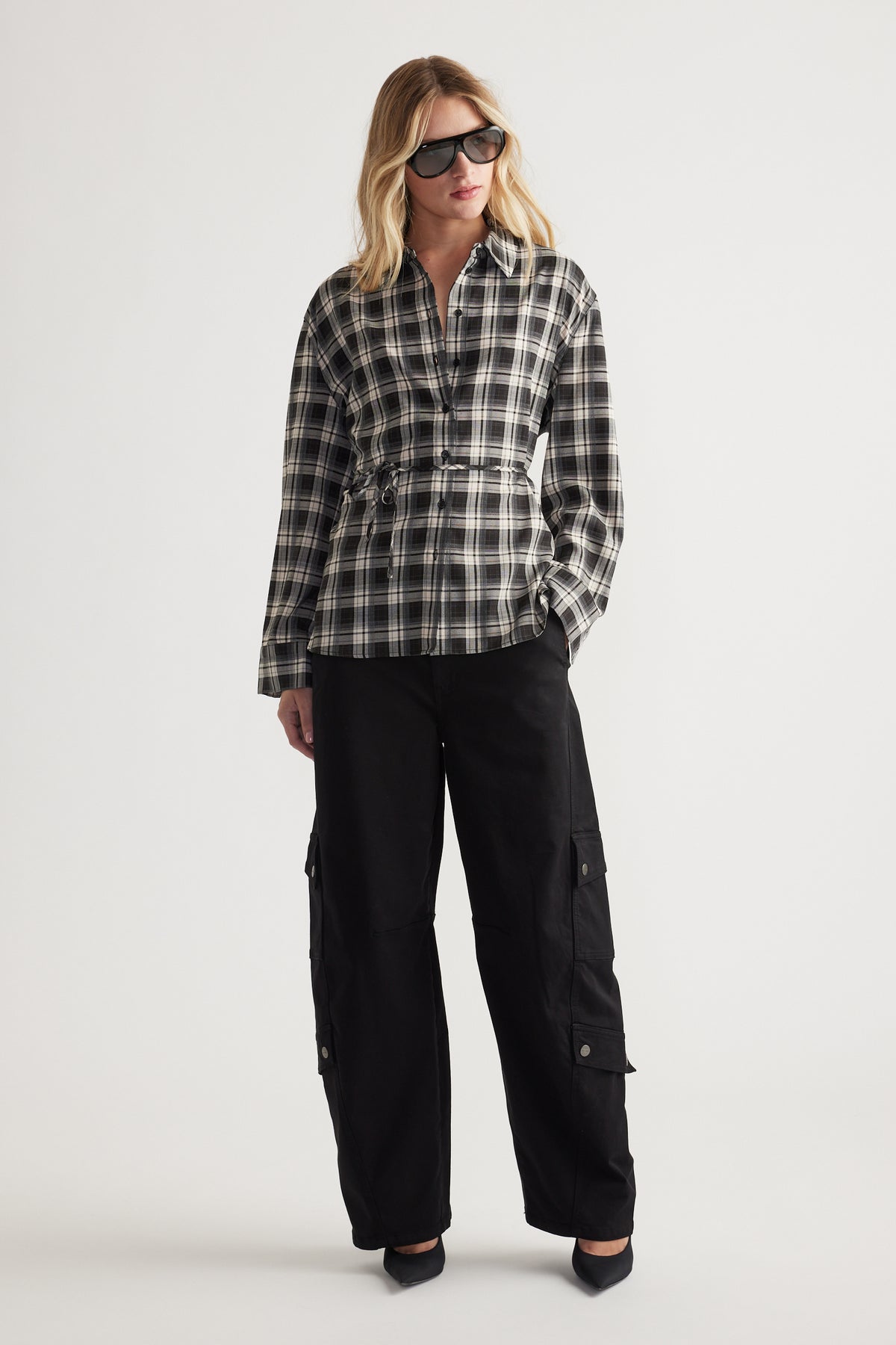 Woman wearing a relaxed fit black and white checked shirt with drawstring waist, paired with loose black cargo pants and sunglasses