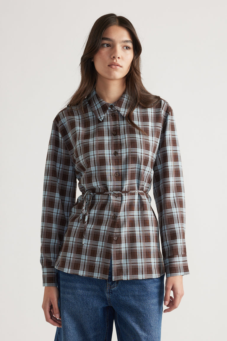 Woman wearing a brown and blue check shirt with a tie waist styled with relaxed denim jeans against a plain background