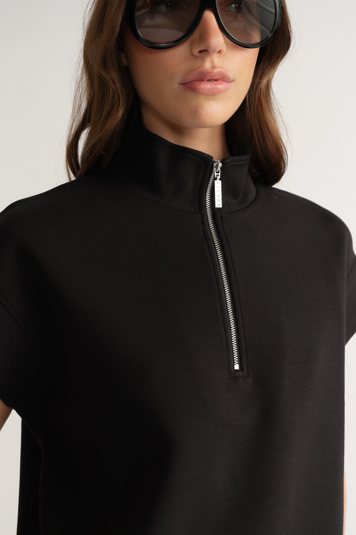 Woman wearing a black short sleeve funnel neck top with a half zip closure and oversized sunglasses
