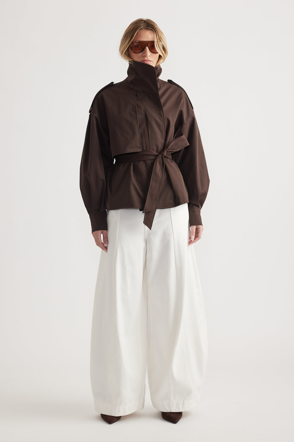 Woman wearing a high-neck dark brown wrap jacket with balloon sleeves paired with wide-leg white trousers and brown shoes