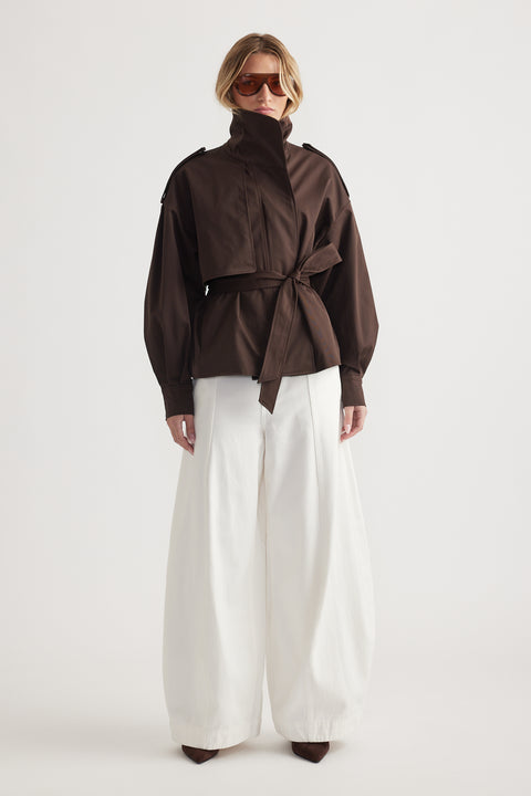 Woman wearing a high-neck dark brown wrap jacket with balloon sleeves paired with wide-leg white trousers and brown shoes