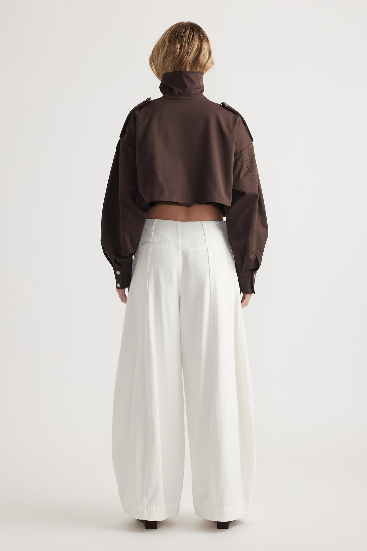 Woman seen from back wearing wide-leg white trousers paired with a cropped brown jacket with a high collar
