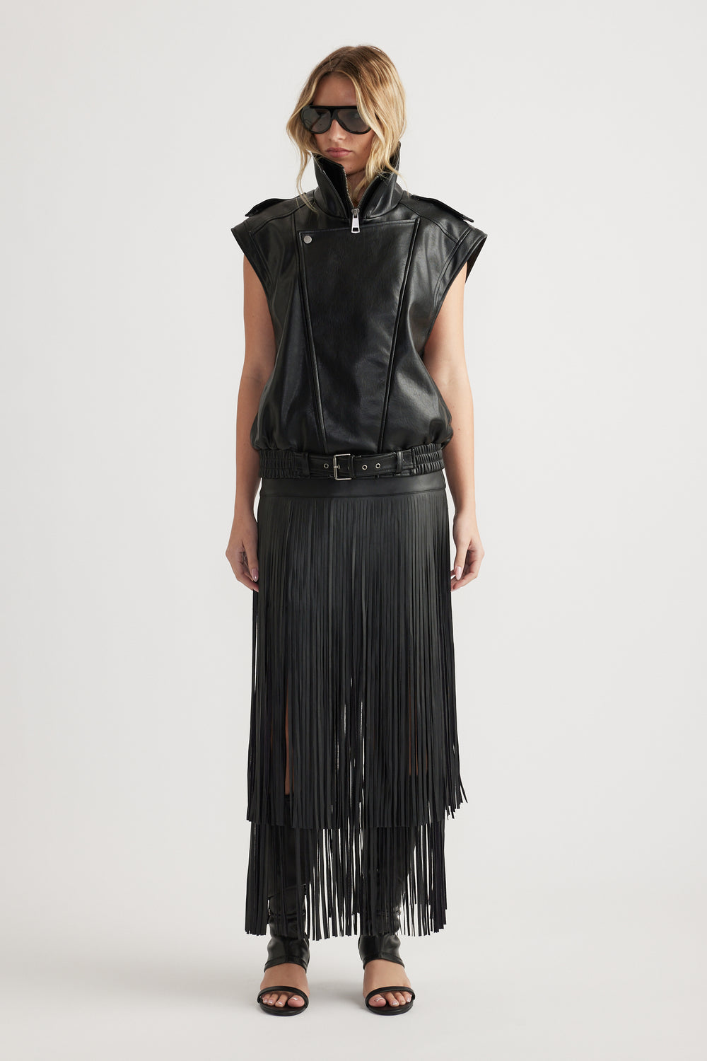 Woman wearing a belted black faux leather vest paired with a long fringed skirt and open-toe heels
